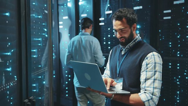 Cheerful IT administrator checking network servers on rack typing data on laptop computer staying in digital server room of data center.