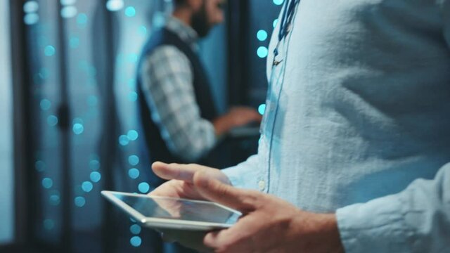 Smart server engineer working in digital network server room. Close-up data center male specialist typing on tablet activating rack server cabinets behind glass panels.