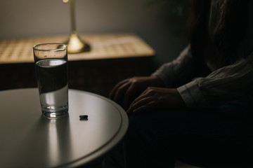 Unhealthy woman is sitting in front of a pill and a glass of water in a dark room, close-up. Girl has insomnia