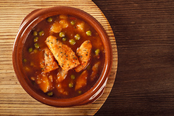Top view of a clay pot with a delicious fish stew with peas and cooked potatoes on a wooden plate on a rustic table. Healthy fish soup. Homemade food. Hot food, freshly made