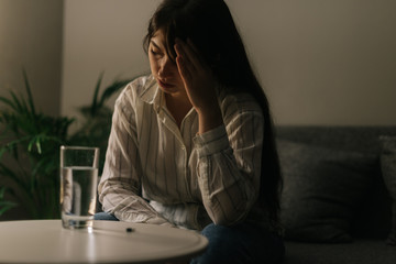 Sick beautiful young woman sitting in front of a table with a tablet and a glass of water in a dark room.
