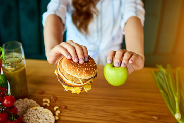 Beautiful young woman decides eating hamburger or apple in kitchen. Cheap junk food vs healthy diet