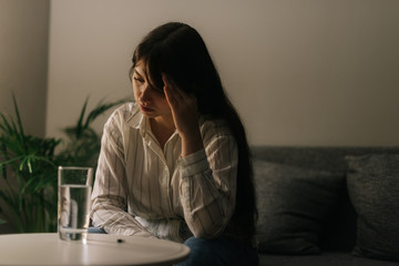 Sick tired beautiful woman sitting in front of a table with a tablet and a glass of water in a dark room.