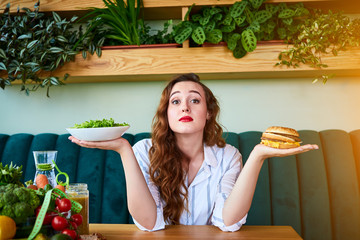 Beautiful young woman decides eating hamburger or fresh salad in kitchen. Cheap junk food vs healthy diet - obrazy, fototapety, plakaty