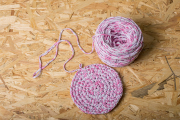 a large skein of thread and a knitted napkin from these threads of white and pink. view from above. all this is on oriented chipboard
