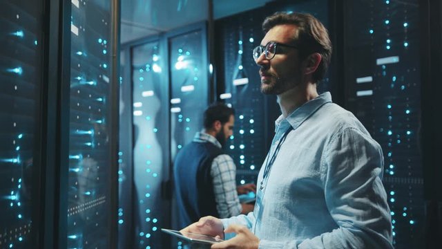 Server technician looking into rack server cabinets updating system with digital tablet. His colleague working on cyber security with laptop computer.