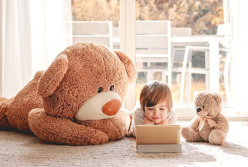 Cute little child watching cartoons on digital tablet device lying on floor with two soft teddy...