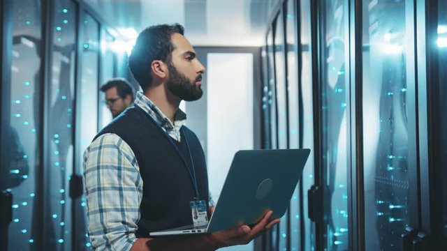 Male administrator inspecting server cabinets on rack typing network data on laptop computer. Team of IT technicians working together in data center server room.