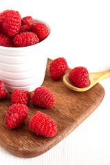 Top view of red raspberries in a white bowl on wooden board, one in a wooden spoon. On white wooden background