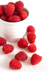 Top view of red raspberries in a white bowl on white wooden board in vertical