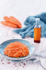 Grated carrot salad on blue plate, peeled carrots, bottle with olive oil, pink peppercorns, blue towel and a fork on the white wooden table. Organic food concept. High key vertical image, copy space