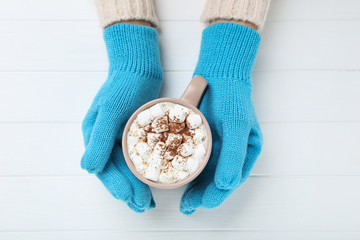Hands in knitted mittens holding cup of hot drink on white wooden table