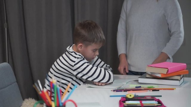 Close-up of an exhausted Caucasian boy sitting at the table as his mother blaming him. Naughty son refusing to study or to do his homework. Bad behaviour, education concept.