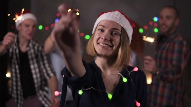 Pretty girl with colorful lights on neck and santa hat posing for camera - smiling, waving her bengal light while her friends dancing on the blurred background in decorated room. Friends celebrating