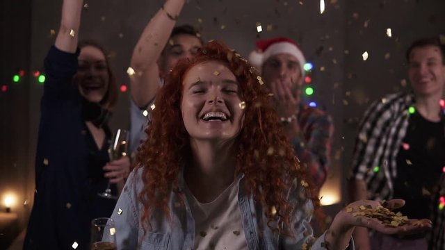 Group of friends celebrating enjoying new years eve party having fun celebration. Portrait of a curly red haired blowing glitter confetti from the hand then after friends throwing confetti on the