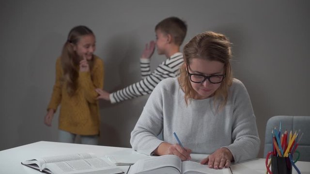 Brother and sister playing at the background as their exhausted mother in eyeglasses sitting at the table and doing homework for her children. Beautiful woman helping her naughty son and daughter.