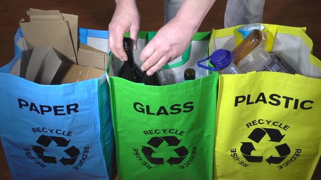Slow motion close POV shot of a man&rsquo;s hand placing two empty glass bottles into an individual portable recycle carrier, with separate carriers for paper and plastic alongside.
