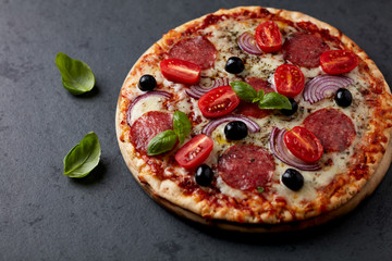 Pizza with salami, mozzarella cheese, cherry tomatoes, black olives, red onion and oregano. Home made food. Concept for a tasty and hearty meal. Black stone background. Copy space