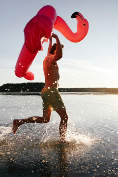 Young man running with flamingo pool float into a lake