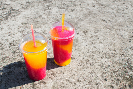 Multi-colored Fruit Cocktail Of Crushed Frozen Ice On A Concrete Background. Place For Text, Layout For Design. Cooling Orange-pink Non-alcoholic Takeaway Sweet Drink In A Plastic Glass.