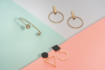 Golden bracelet and two golden geometric earrings pairs on pastel colors background pink and blue