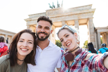 Fototapeta premium Portrait of three happy friends taking selfie with cell phone in front of Brandenburger Tor, Berlin, Germany