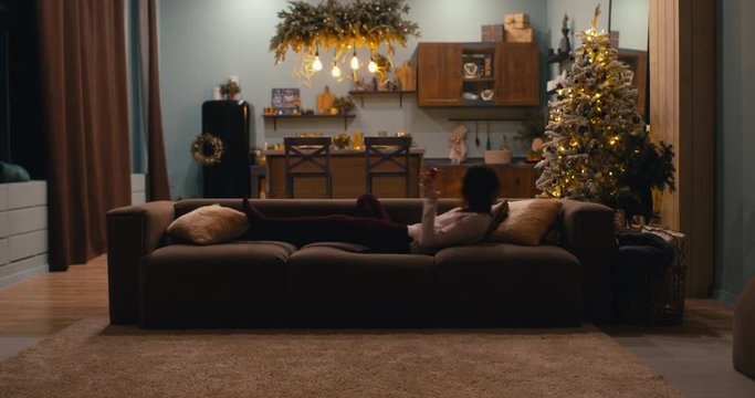 WIDE Caucasian female jumping to a sofa and checking her phone at home during Christmas holidays, decorated apartment interior. 4K UHD RAW graded footage