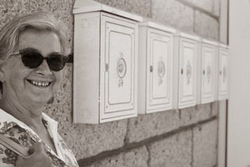 Attractive senior caucasian woman of 70 years smiling and standing close to the mailboxes of house. A sunny day