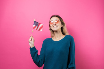 Happy young woman heart-shaped glasses holding American flag against a studio pink background