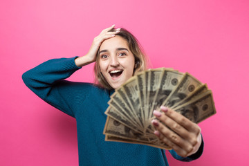 Closeup of young beautiful woman in a blue sweater in glasses with american dollars money in hand over pink background. Expresses emotions and gestures of joy.