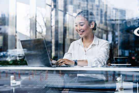 Young Asian Woman Drinking Coffee And Using Laptop At A Cafe With Airpods In Ears