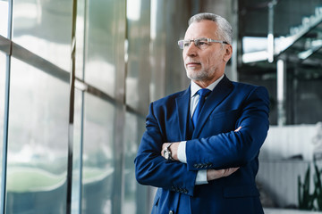 Side view of senior businessman in suit standing in office lobby and looking away through window