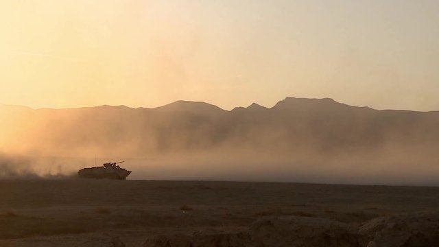 Military heavy combat vehicle rides through the desert raising the dust, the evening sun in the mountains and on the plain.