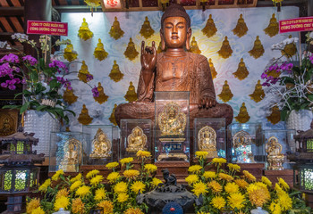 Da Nang, Vietnam - March 10, 2019: Chua An Long Chinese Buddhist Temple. Brown wooden central Buddha statue on main altar. Yellow flowers and golden ornaments in front. Small glass buddhas as backdrop