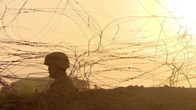 A peacekeeper soldier in a helmet and with a machine gun guarding a checkpoint in the desert, fences and barbed wire, sunrise, silhouette lighting.