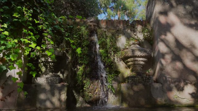 This slow motion serene video shows an ancient fountain, overgrown with lush greenery, with water flowing from mouth at Parc del Laberint d'Horta in Barcelona, Spain on a sunny day.