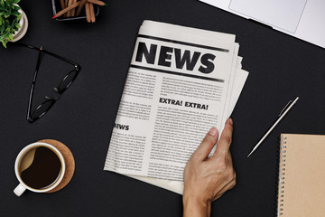 Hand holding Business Newspaper and office supplies with notebook, hot coffee cup, glasses, books and accessories on black desk topview, Daily Newspaper mockup concept