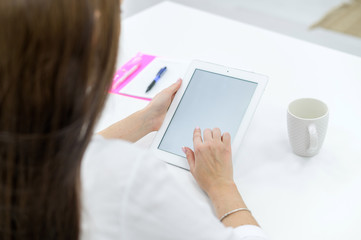 A young woman in a white shirt and jeans sits at a table with a tablet in her hands. A female office manager performs work on a modern device. The view from behind.
