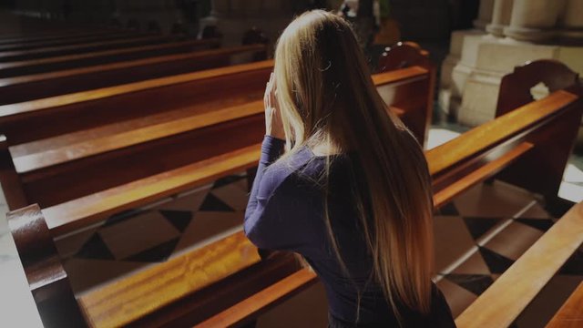 This video shows a young caucasian woman kneeling and praying at beautiful catholic church pew, rear view with sun beaming down.