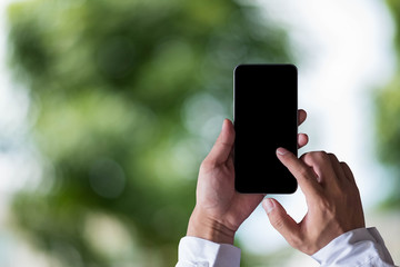 Hand holding white mobile phone with blank white screen in forest in the morning.