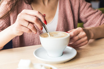 Attractive young woman sits at a table in a cafe with a cup of coffee and mixes it with a spoon