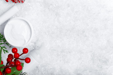 Facial skincare products flat lay on snow background. Protective lip balm, cream jar, balm bottle mockups with mistletoe berries decorative backdrop. Cold season cosmetic product advert with copyspace