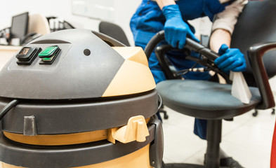 Young man in workwear and rubber gloves cleans the office chair with professional equipment.