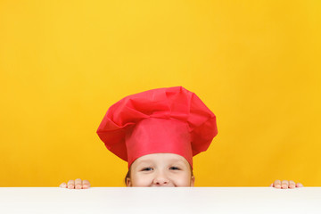 Funny little girl chef on a yellow background. The child is hiding and looking out from under the table