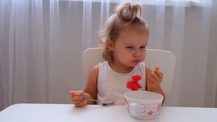 Infant girl eating baby food at a small childrens table on a white background. Nutrition and feeding of children.