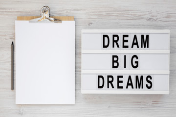 'Dream big dreams' words on a lightbox, clipboard with blank sheet of paper on a white wooden surface, top view. Overhead, from above, flat lay.