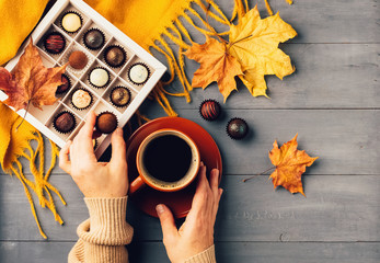 Woman's hands holding a cup of hot black coffee orange scarf box of chocolates autumn background. Top view flat layout.