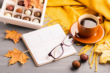 A cup of hot black coffee notepad diary glasses orange scarf box of chocolates autumn background. Top view flat layout.