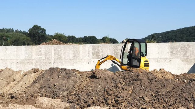 Yellow mini excavator digs a hole at a construction site. A small excavator pulls the earth out of the pit.