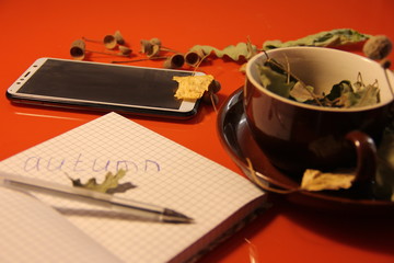 Great autumn background. in a brown mug are dry oak leaves and acorns. Nearby lies a phone - a smartphone and a notepad